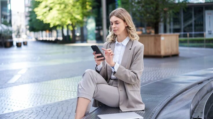 Uma mulher de negócios sentada em uma saliência e usando o celular.